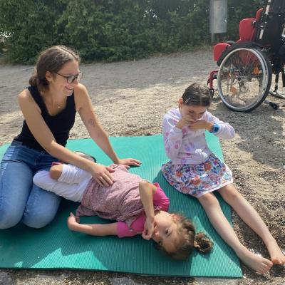 Zwei Mädchen sind auf einer Matte auf dem Boden positioniert. Eine Liegend, eine Sitzend. Daneben sitzt eine Lehrerin. Beide sind direkt am Bachlauf und können das Wasser mit ihrer Hand bzw. mit dem Füßen spüren. 