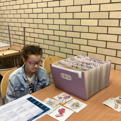 Eine Person sitzt an einem Tisch. Vor ihr liegt ein Blatt. Daneben liegen mehrere Bildkarten mit Lebensmitteln darauf. Daneben steht ein Fächerordner. 