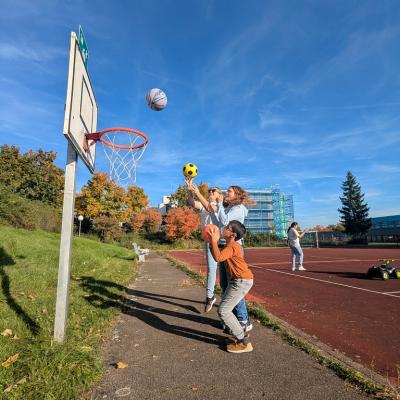 Man sieht 3 Personen in einer Reihe. Ganz hinten steht eine Frau. In der Mitte ein junges Mädchen. Ganz vorne ein Junge. Alle drei Werfen einen Ball in den Basketballkorb vor ihnen. 