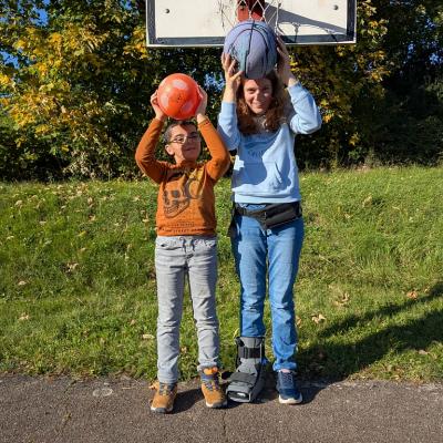 Ein junges Mädchen und ein Junge stehen nebeneinader. Sie lachen und strecken ihre Hände in Luft. In den Händen haben sie ein Ball. Im Hintergrund steht ein Basketballkorb