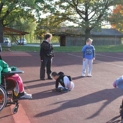 Es stehen Menschen und zwei RollstuhlfahrerInnen im Kreis auf dem Sportplatz, in der mitte sitzt ein Junge auf dem Boden.