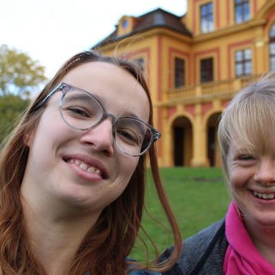 Zwei junge Frauen lachen in die Kamera, im Hintergrund sieht man das Schloss im Favorite Park