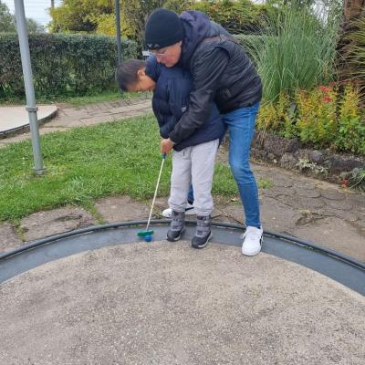 Eine Frau und ein Junge sind auf einer Minigolf-Bahn.Der Junge steht vorne. Die Frau steht hinter ihm.Sie hilft dem Jungen beim Spielen 