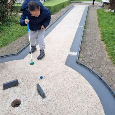 Ein Junge steht auf einer Minigolf- Bahn.Er versucht den Ball in das Loch zu schlagen. 