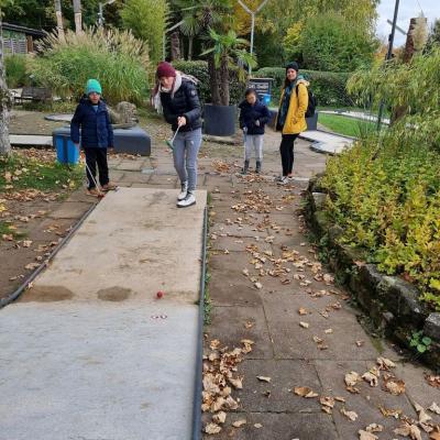 Man sieht vier Personen.Vorne steht ein Mädchen.Das Mädchen schlägt einen Minigolf-Ball.Der Ball rollt die Bahn hoch. Die Bahn hat einen kleinen Hügel.