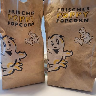 Man sieht zwei Braune Tüten mit dem Schriftzug: Frisches Poppy Popcorn.  Daneben und darunter sieht man kleine Popcorn-Männchen. 