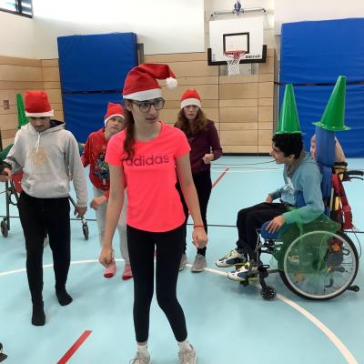 Auf dem Foto sind Schülerinnen und Schüler zu sehen die in einer Sporthalle joggen und dabei Weihnachtsmützen tragen. Ein Schüler im Rollstuhl ist mit einem grünen Hütchen und grünen Tüchern als Tanne verkl
