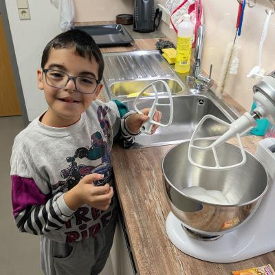 Ein Junge steht vor der Küchenmaschine und macht Zutaten hinein, lacht in die Kamera