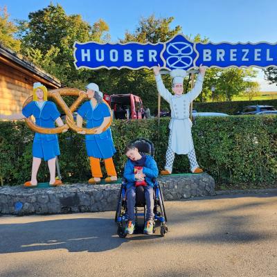 Schüler im blauen Rollstuhl steht vor drei Figuren vorm Brezel Huober Laden. 