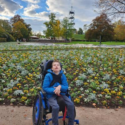 Schüler sitz in blau, schwarzen Rollstuhl, mit blauer Jacke und schaut in die Camara, im Hintergrund sind gelbe, grüne pflanzen im Beet