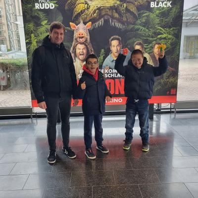 Es stehen von links nach rechts in einer Reihe ein Mann und 2 Jungen vor einem Filmplakat im Kino.