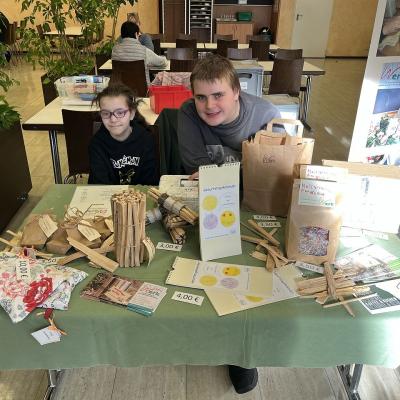 Auf diesem Bild sitzen zwei Schüler am Verkaufsstand. Die Schüler verkaufen Feuerbälle, Samenbomben, Dinkelkissen, Spächtele to go und Geburtstagskalender.  Zwei Schüler freuen sich auf Kunden.   