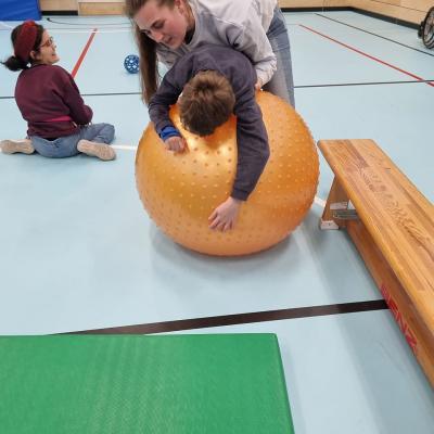 Ein Junge liegt mit dem Bauch auf einem großen Pezziball. Über ihm steht eine Frau und hält ihn fest. 