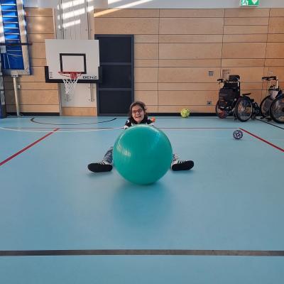 Ein Mädchen sitz auf dem Boden der Turnhalle. Sie spielt mit einem türkisfarbenen Pezziball. 