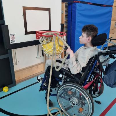 Eine Junge im Rollstuhl sitzt vor einem Basketballkorb. Er hält ein Seil mit der Hand fest, das Seil ist im Basketballkorb 