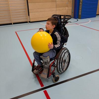 Ein Junge im Rolstuhl spielt mit einem großen gelben Gymnastikball