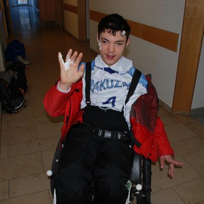 Maik steht mit seinem Rollstuhl im Gang vor seinem Zimmer. Er hat seine Skiklamotten an und Sonnencreme an der Hand, an der Backe und in den Haaren.