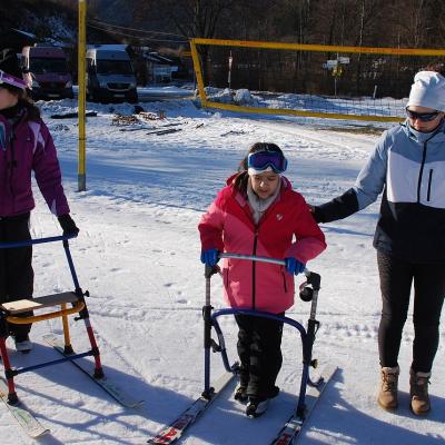 Marija, Ghofran und Frau Fecke stehen im Schnee. Marija und Ghofran halten sich je an einem Skilator fest. Im Hintergrund sieht man ein gelbes Volleyballnetz und dahinter die geparkten Busse.
