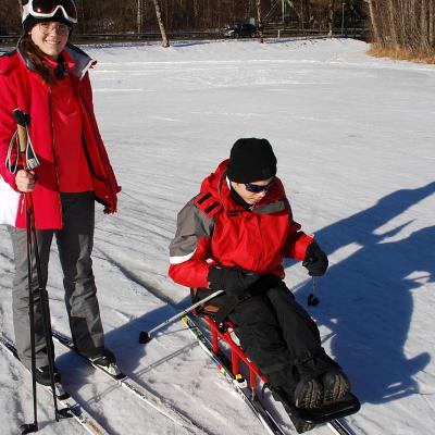 Lena steht mit Ski im Schnee und lächelt in die Kamera. Daneben sitzt Maik im Skischlitten und stochert sich voran.