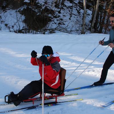 Maik stochert im Skischlitten in der Loipe. Herr Kling läuft dahinter und schiebt den Schlitten mit seinen Skistöcken.