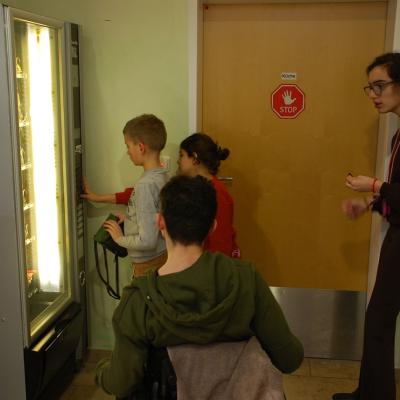 Luca, Sophia, Maik und Marija stehen mit Geld in der Hand vor einem Snack-Automat. Luca drückt gerade auf eine Taste, um etwas herauszulassen.
