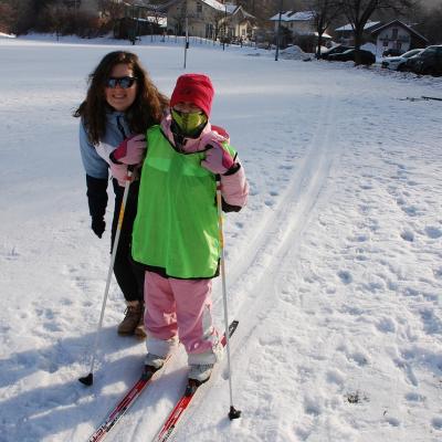 Sophia fährt in der Loipe. Ihr Gesicht ist gut verhüllt. Hinter ihr lächelt Frau Fecke in die Kamera. Die Sonne wirft ihre Schatten neben sie in den Schnee.