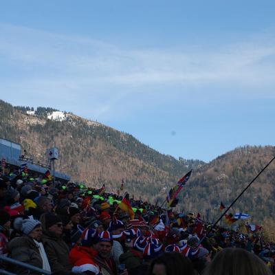 Man sieht eine gefüllte Tribüne. Sie haben verschiedene Länderfarben in den Klamotten. Im Hintergrund sieht man Berge und blauen Himmel.