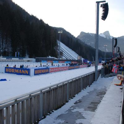 Man sieht links die Schießanlage. Dahinter ist eine Sprungschanze und bewaldeter Hügel. Rechst davon ist die Zielgerade und daneben die Zuschauertribüne.