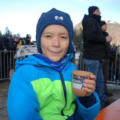 Luca sitzt auf einer Bierbank, hält einen Becher in der Hand und grinst in die Kamera.