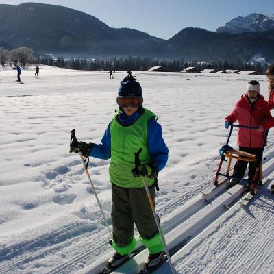 Luca läuft in der Loipe. Dahinter läuft Ghofran am Skilator und neben ihr Lena. Im Hintergrund sieht man bewaldete Berge und blauen Himmel.