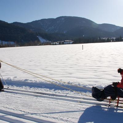 Maik fährt im Skischlitten in der Loipe und wird von Herrn Kling gezogen, indem dieser ein Seil umgebunden hat und ihn so zieht. Dahinter Berge und Himmel.