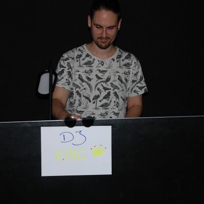 Herr Kling steht hinter dem DJ-Pult. Daran hängt ein Schild: 