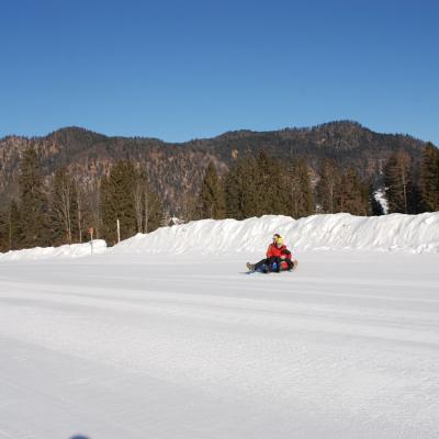 Maik und Herr Kling sitzen zusammen in einem Reifen und fahren auf der Geraden nach dem Hang. Dahinter sieht man Wald, Berge und Himmel.