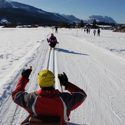 Man sieht Maik von hinten in der Loipe stochern. Vor ihm fährt Marija. Neben ihnen ist die Skating-Strecke. Im Hintergrund bewaldete Berge und Himmel