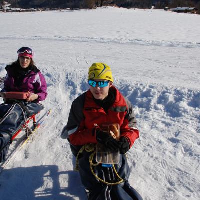 Marija und Maik sitzen in ihren Skischiltten und haben ihre Vesperboxen in der Hand. Daneben steht eine graue Kiste und ein Holzschlitten.