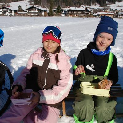Sophia und Luca sitzen auf einem Holzschlitten und haben ihr Vesper auf dem Schoß. Rechts sitzt noch eine Person. Links sieht man einen Rolli.