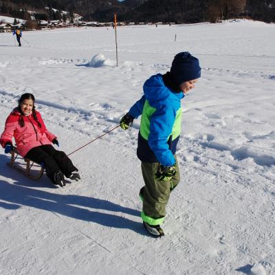 Luca zieht den Holzschlitten über den Schneeweg, auf dem Ghofran sitzt. Dahinter sieht man Schnee, Häuser und Wald.