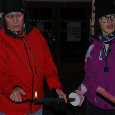 Marija hält eine schwarze Fackel, die von Frau Gauch angezündet wird. Es ist offensichtlich Nacht. Beide tragen warme Kleidung und sind im Freien.