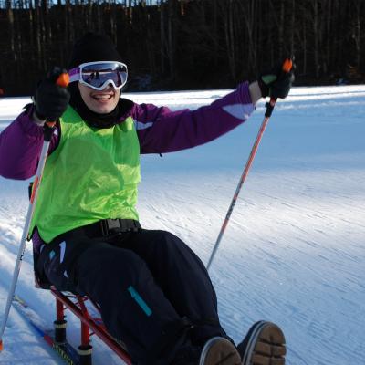 Marija stochert im Skischlitten in der Loipe. Sie lacht und hat sichtlich Spaß. Dahinter sieht man die Loipe und dunkle Bäume.