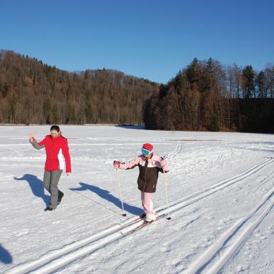 Sophia läuft in der Loipe. Lena feuert sie daneben mit einer Rätsche an. Dahinter sieht man Schnee, dunkle Bäume und blauen Himmel. Rechts steht Ghofran.