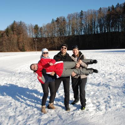 Frau Fecke, Herr Kling und Frau Gauch tragen Lena, die horizontal über deren Arme liegt. Sie zeigt Daumen hoch. Alle lächeln in die Kamera. Dahinter Bäume.