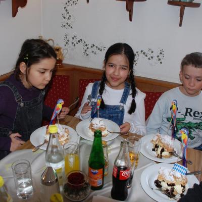 Sophia, Ghofran und Luca sitzen auf einer Eckbank am Tisch. Alle drei haben Windbeutel vor sich, in denen eine Papier-Papagei steckt.