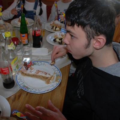 Maik sitzt am Tisch, vor sich ein Stück Apfelkuchen. Auf dem Tisch stehen andere Speisen und Getränke. Maik isst seinen Kuchen mit einer Gabel.