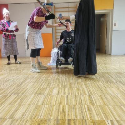 Berta hält Maik den Langlaufski in den Mund. Der Hohepriester schüttet Wasser darüber in seinen Mund. Leni hält daneben seine Urkunde.