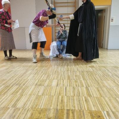 Berta hält Sophia den Langlaufski in den Mund. Der Hohepriester schüttet Wasser darüber in ihren Mund. Leni hält daneben ihre Urkunde.