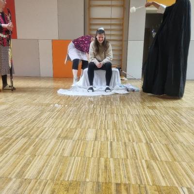 Lena sitzt auf dem Thron und hat Schnee auf dem Kopf. Sie wird vom Hohepriester nassgespritzt. Berta beugt sich hinter sie. Leni hält den Langlaufski