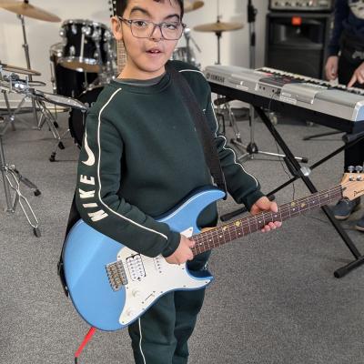 ein Junge hat einen Bass/Gitarre um und lacht in die Kamera