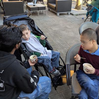 Ein junger Mann sitzt in einem Buggy, ein junger Mann und ein Mann sitzen auf Stühlen. Alle essen ein Eis. 