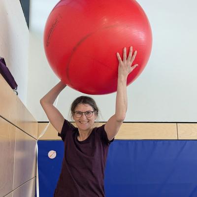 Eine Frau hält einen großen roten Gymnasikball hoch und lacht in die Kamera