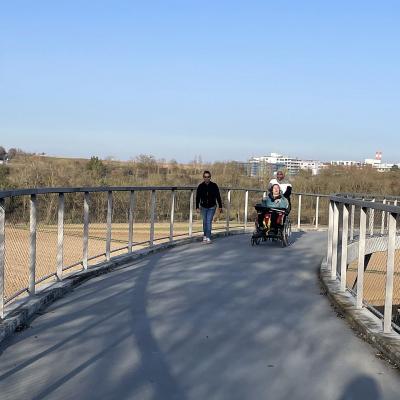 Eine Frau schiebt eine junge Frau im Rollstuhl, eine andere Frau läuft nebenher. Sie laufen über eine Brücke